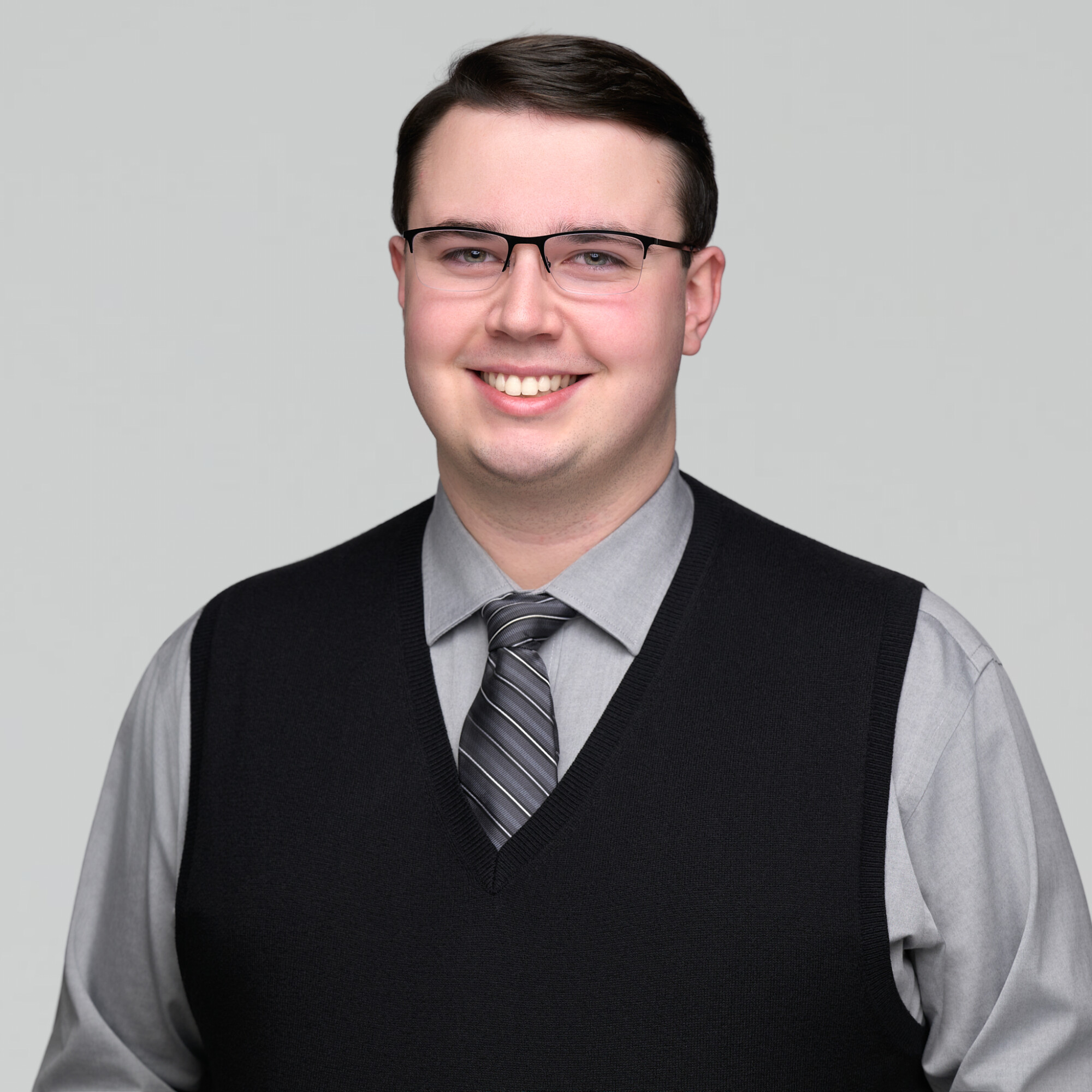 Professional headshot of a smiling man wearing glasses, a gray dress shirt, striped tie, and black sweater vest against a light gray background.